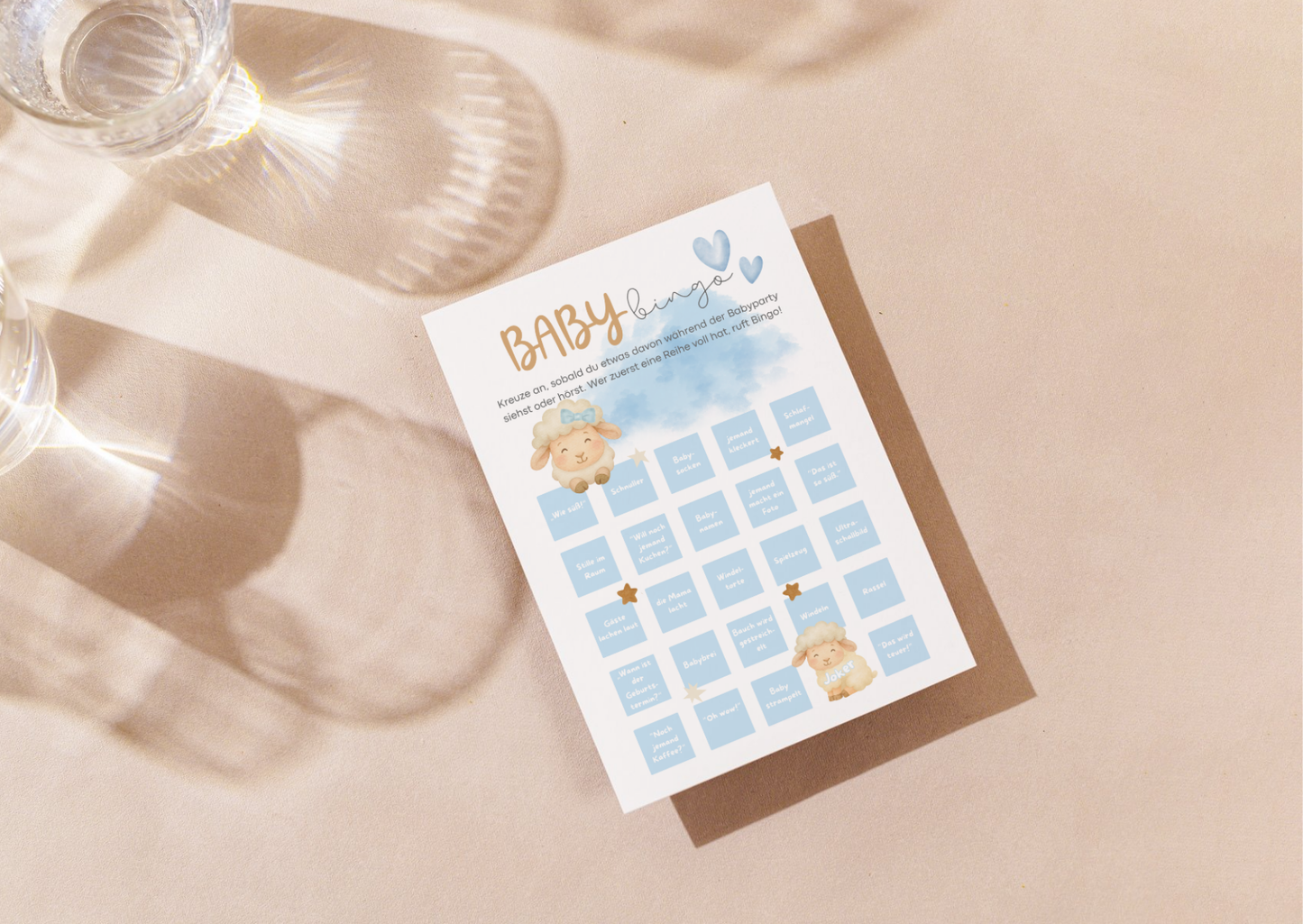 Baby Bingo Karte für Babyparty im Boho Stil, blaues Design mit Lammmotiv auf hellem Tisch mit Glasreflexen