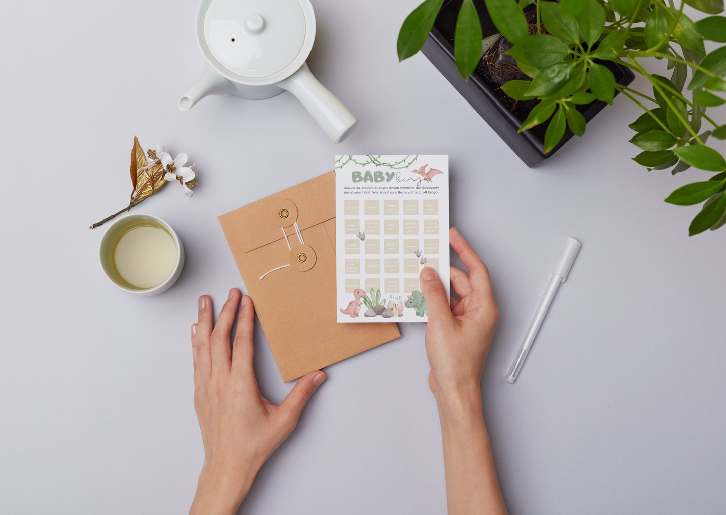 Person hält Baby Bingo Karte mit Dinosaurier-Motiv im Boho Stil, nachhaltiges Partyspiel für Babyparty auf hellem Tisch mit Pflanzen