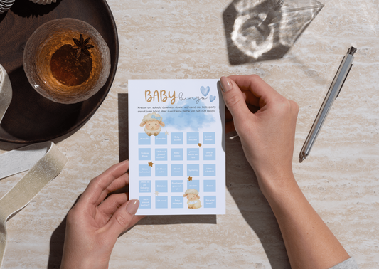 Person hält Baby Bingo Karte im Boho Stil auf Holztisch mit Kaffee, süßes Babyparty Spiel mit Lammmotiv in Blau